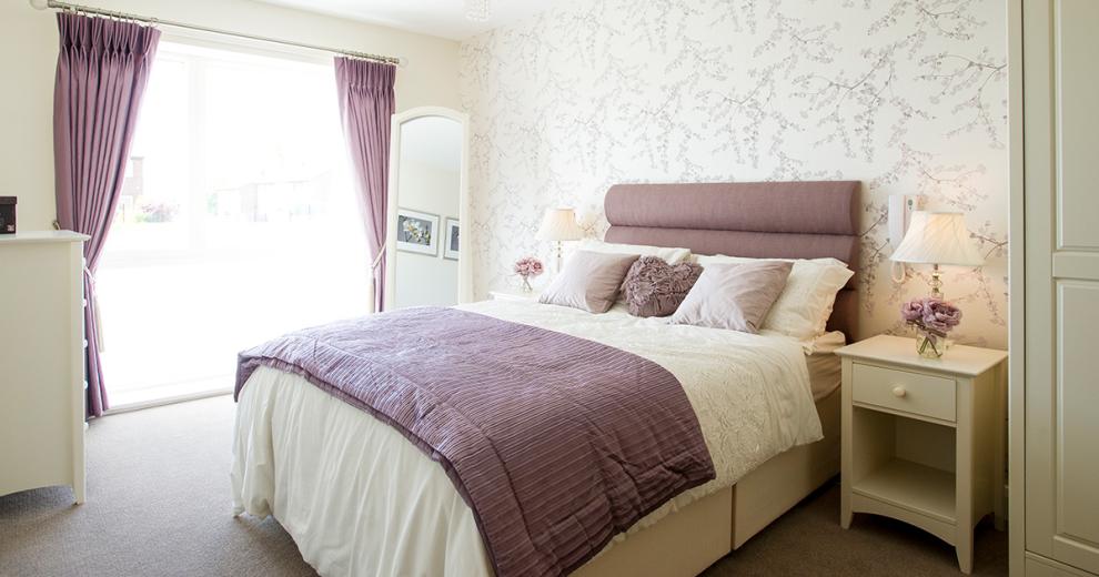 Peaceful double bedroom, tastefully decorated with delicate lilac blossom flowers on a cream background. Generous sized window allowing an abundance of light into the room. White free-standing mirror and matching white furniture set consisting of two bedside tables, a chest of draws and wardrobe add functionality with a clean design.