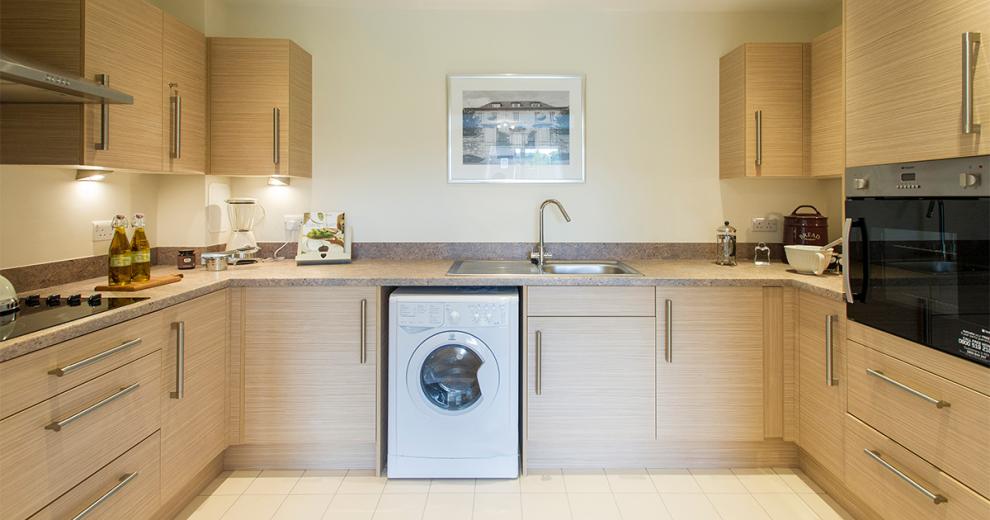 Well sized modern kitchen featuring a wall mounted oven, opposite a ceramic hob, a washing machine and numerous cabinets and draws in a light wooden finish and matted chrome handles surrounding the space.