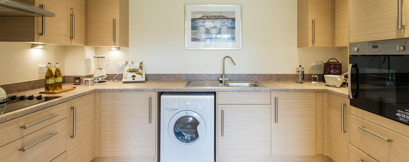 Well sized modern kitchen featuring a wall mounted oven, opposite a ceramic hob, a washing machine and numerous cabinets and draws in a light wooden finish and matted chrome handles surrounding the space.