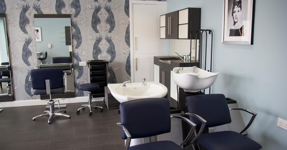 Roomy onsite hair salon, using a classic muted colour palette throughout. Navy and silver hairdressing chairs pop against the white basins or in the reflections of the large mirrors.