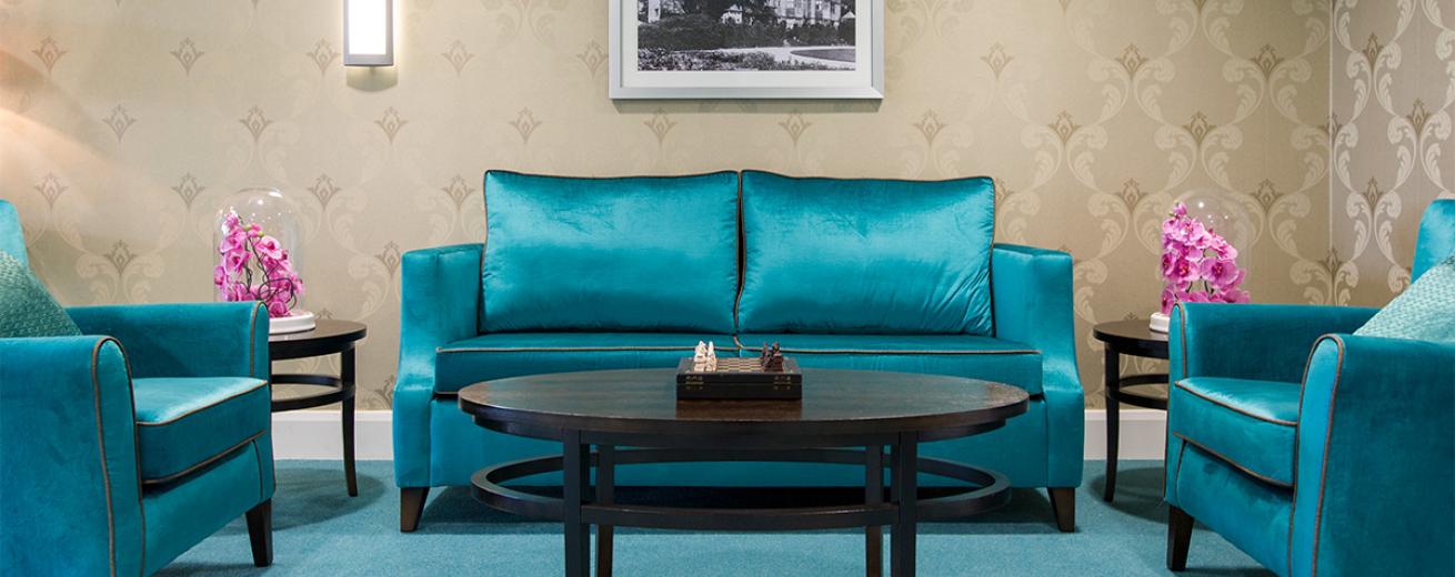 Bold teal seating area with plump crushed velvet two seater sofa and two arm chairs arranged around a large oval coffee table with two smaller circular tables placed either side of the sofa. The teal carpet mates the seating décor and contrasts with the earth toned, fleur de lis wallpaper.