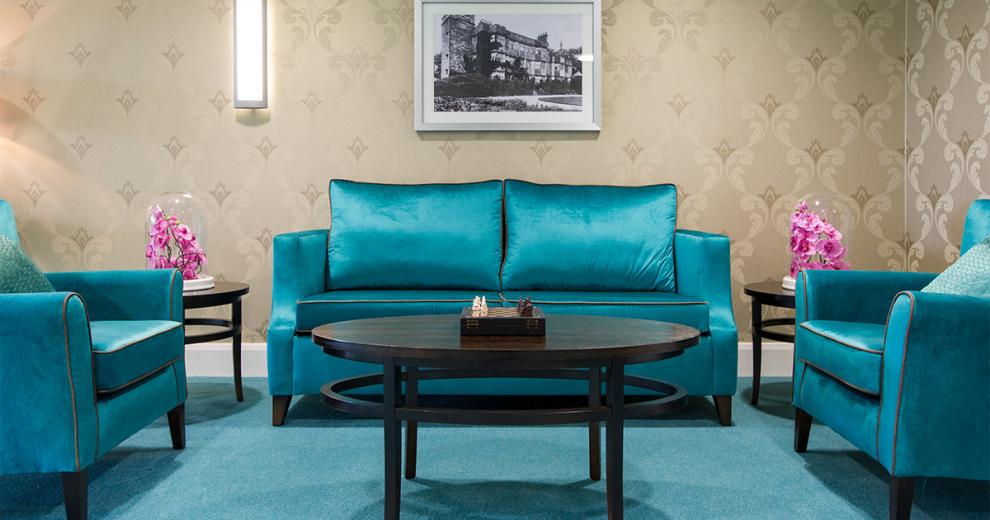 Bold teal seating area with plump crushed velvet two seater sofa and two arm chairs arranged around a large oval coffee table with two smaller circular tables placed either side of the sofa. The teal carpet mates the seating décor and contrasts with the earth toned, fleur de lis wallpaper.