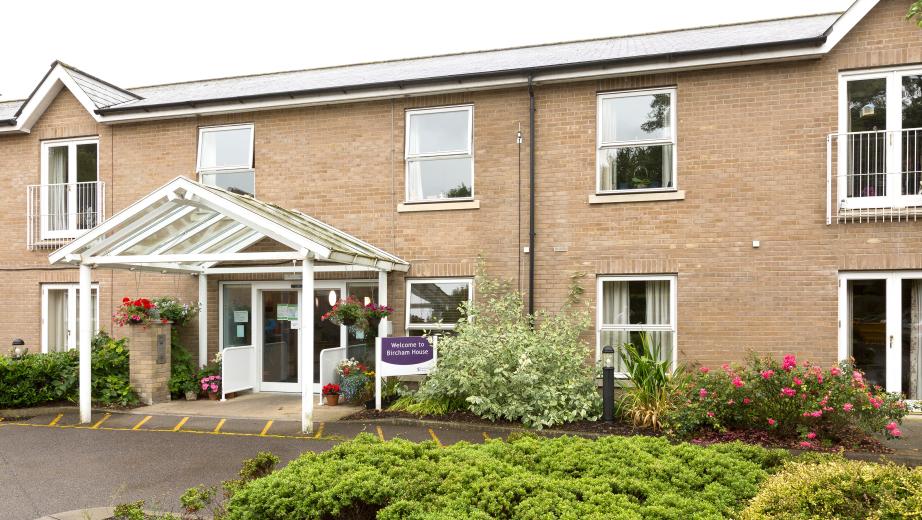 Two storey sandy brick-built building with juliet balconies wrapped around the french doors of the first storey. A mixture of mature shrubbery and plants decorate the perimeter of the building. The entranceway has no step access and is covered by an exposed porch, leading out onto the driveway.