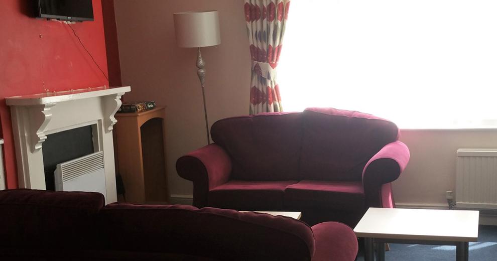 Cosy, seating area consisting of two facing two-seater burgundy sofas, an electric fire surrounded by a simple wood fireplace.