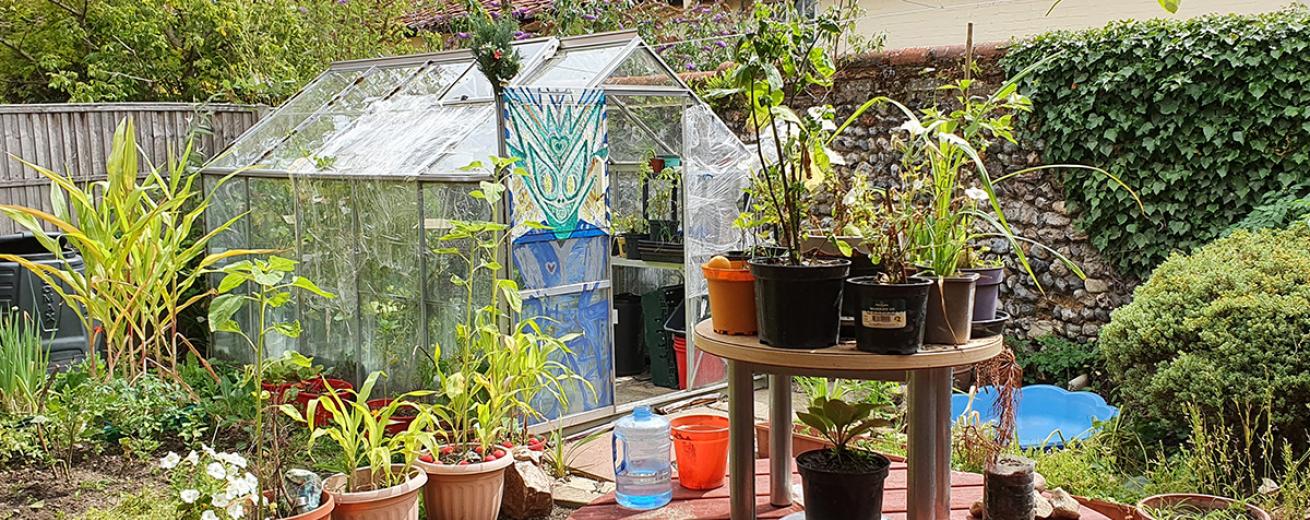 Sizable greenhouse with illustrative artwork, next to a stone separating wall and luscious mixture of plants. Circular table doubling as a potting plant station.