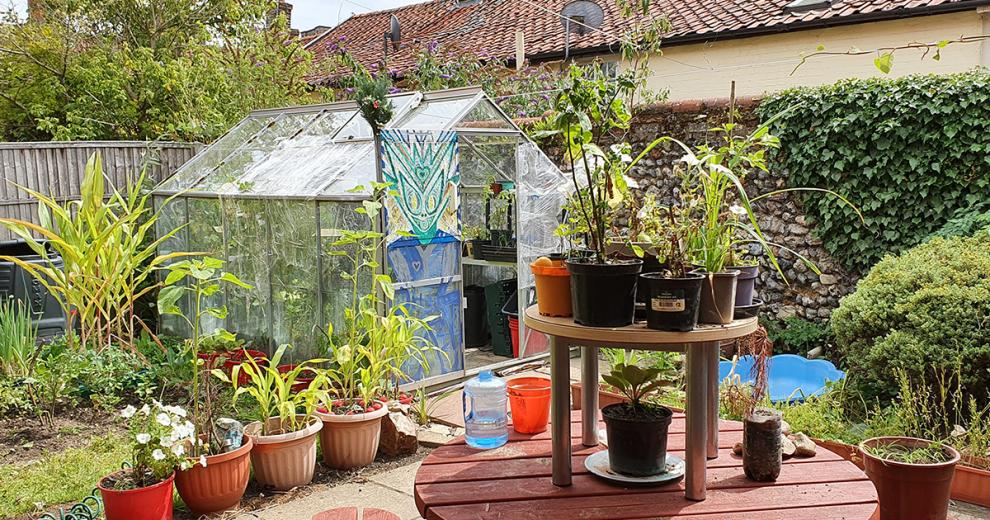 Sizable greenhouse with illustrative artwork, next to a stone separating wall and luscious mixture of plants. Circular table doubling as a potting plant station.