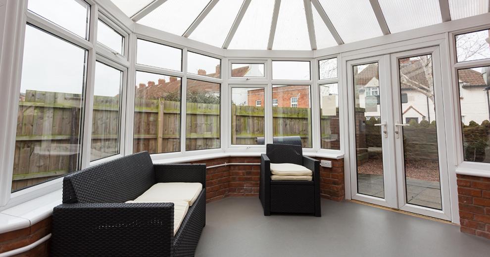 Relaxing angled conservatory room with wicker furniture set, allowing a flood of light from any time of day and French door access to a garden area.