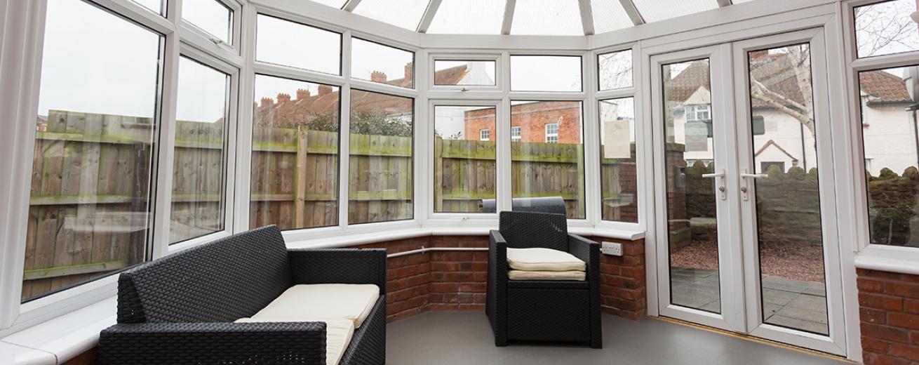 Relaxing angled conservatory room with wicker furniture set, allowing a flood of light from any time of day and French door access to a garden area.
