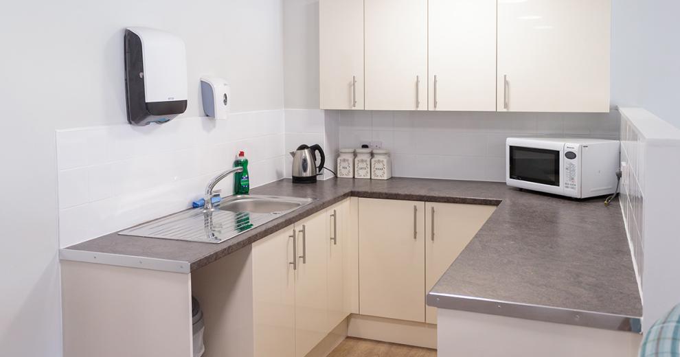 A compact, white communal kitchen area fitted with a host of off-white cabinets. The grey speckled work tops house a microwave, kettle, tea making facilities and an industrial style sink.