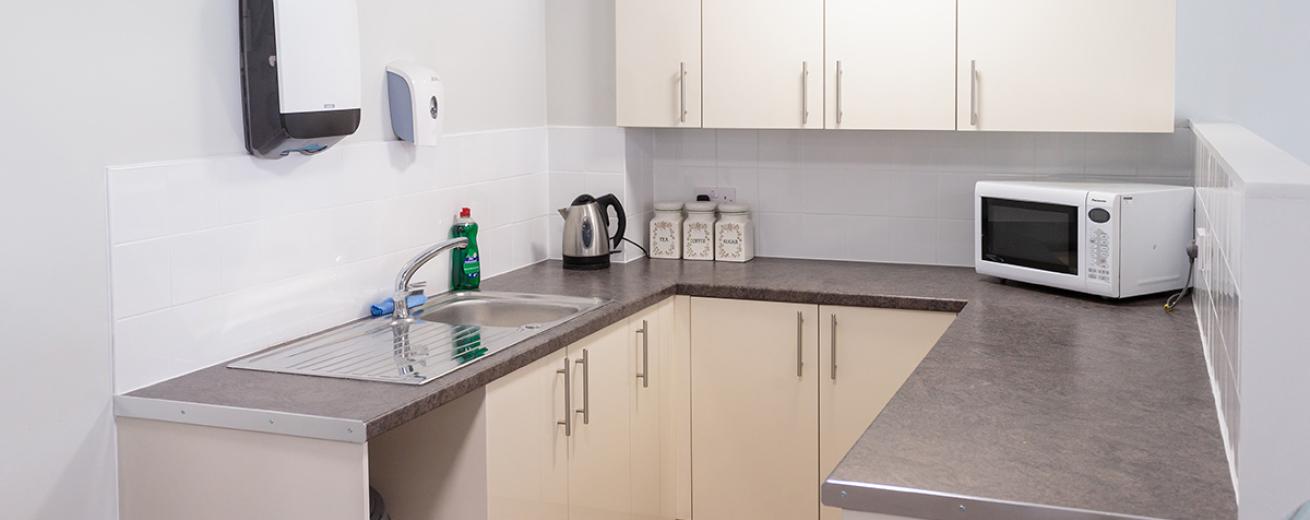 A compact, white communal kitchen area fitted with a host of off-white cabinets. The grey speckled work tops house a microwave, kettle, tea making facilities and an industrial style sink.
