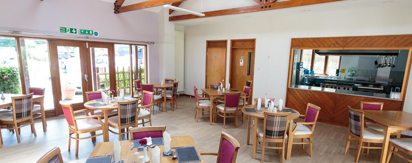 A roomy dining area with access and views into the communal garden An array of four person light wooden dining tables sets are located in front of the restaurant serving hatch.