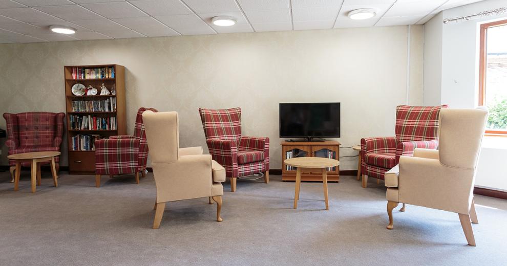 An informal seating area with a mixture of tartan patterned and cream covered chesterfield styled armchairs, located around a television and bookshelf all hosting a small wooden coffee table.