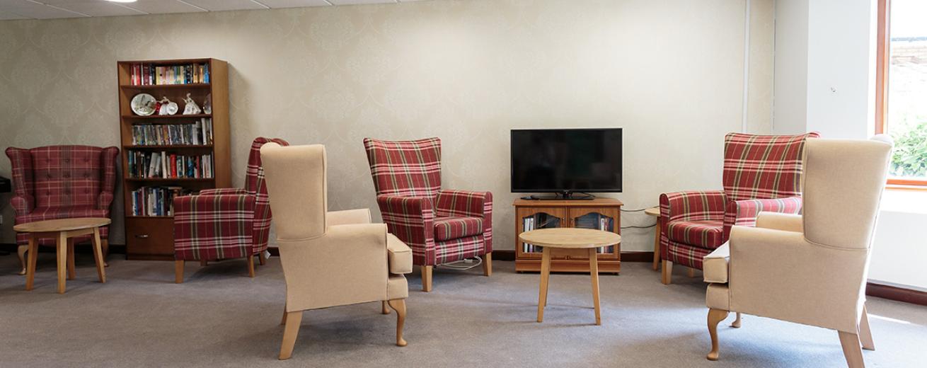 An informal seating area with a mixture of tartan patterned and cream covered chesterfield styled armchairs, located around a television and bookshelf all hosting a small wooden coffee table.