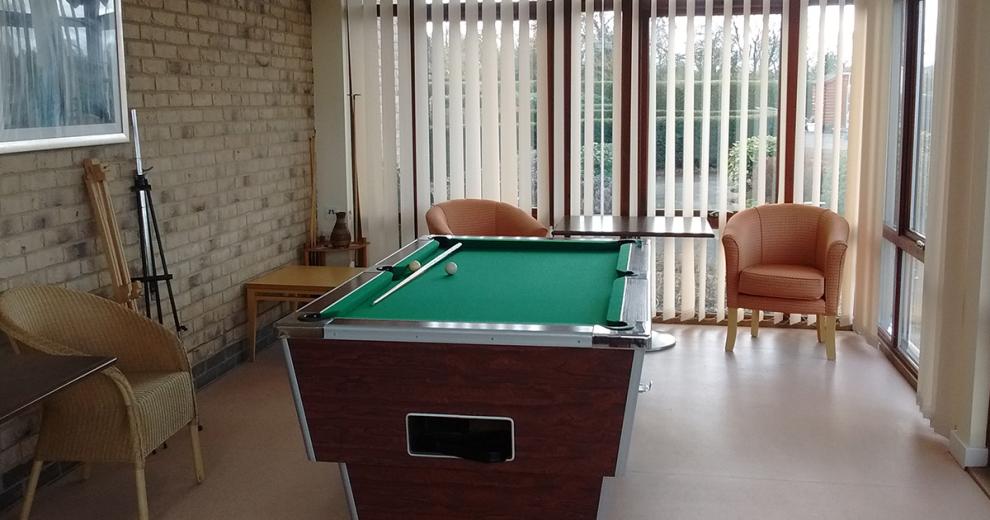A conservatory room with a wooden and chrome finished snooker table located in the centre, small leather armchairs are placed around the table.