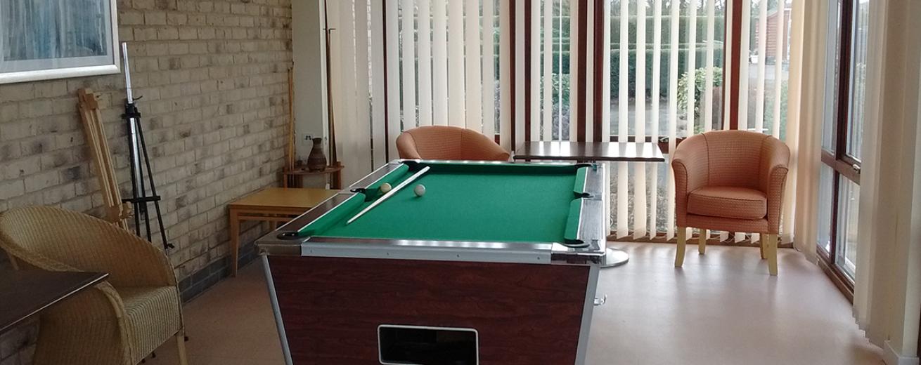 A conservatory room with a wooden and chrome finished snooker table located in the centre, small leather armchairs are placed around the table.