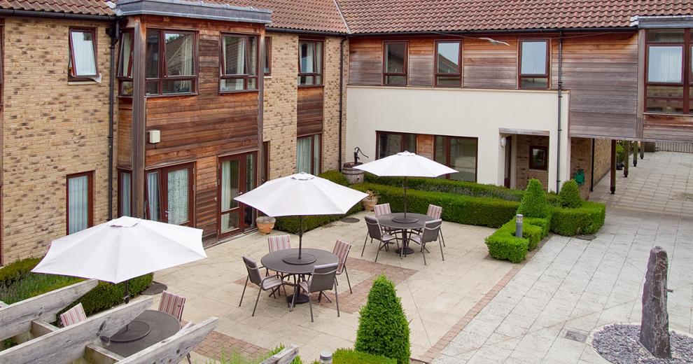 Patio area consisting of 3 outdoor seating sets with umbrella coverings, surrounded by small hedges.