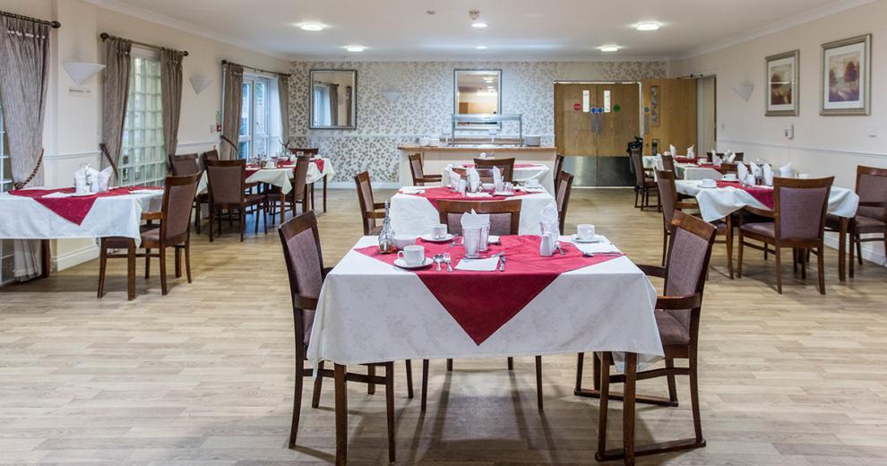 Spacious, light dining area with white and red linen dressed dining tables, set with teacups, saucers, cutlery, and napkins. Set in a floral silver and white patterned decorated room, with matching silver hanging picture frames. As well as silver crushed velvet curtains in front of the large windows and french doors.