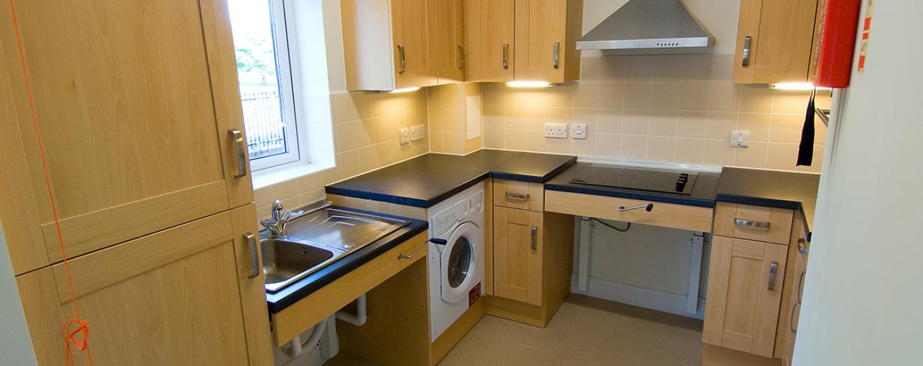 A fully accessible fitted kitchen, with adjustable height sink and electric hob. The light wooden cabinetry and under cabinet lighting gives the kitchen a warming glow.