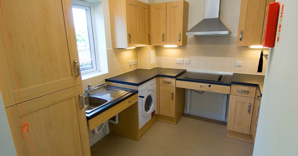 A fully accessible fitted kitchen, with adjustable height sink and electric hob. The light wooden cabinetry and under cabinet lighting gives the kitchen a warming glow.