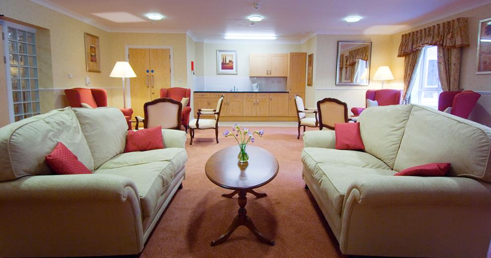 A compact seating area featuring to facing two-seater cream fabric sofas with an oval dark brown wooden table. Located behind are two clusters of seating including chesterfield arms chairs and wooden chairs with arm rests upholstered in the same cream fabric as the sofas. Both clusters of seating house a floor lamp for extra lighting. A kitchenette is built into the alcove of the room.