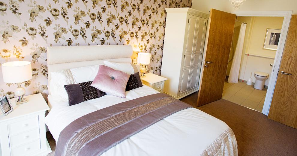 A sophisticated ensuite double bedroom. With a tasteful green and white floral peony wallpaper to create a feature wall. The whitewashed bedside tables and wardrobe contrast well with the darker carpets and dark wooden doors.
