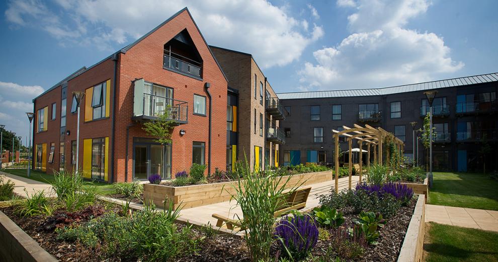 The neatly laid out garden area provides a peaceful and calming atmosphere, with white slab paved walkways bordered by raised wooden plant beds, planted with an assortment of vegetation. Newly planted trees sit behind the raised planters on the well-kept grass.