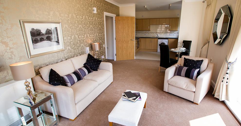 A beautifully presented open plan living area, the largest wall has a subtle silver fleur de lis wallpaper, in the centre is a large black and white bridge photograph. Underneath is a cream two seater sofa and two glass coffee tables with matching lamps either side. A single arm chair is opposite the sofa next to a window allowing a lot of natural light, a cream leather foot rest is placed centrally. A small two person dining table tucks neatly between the living area and kitchen.