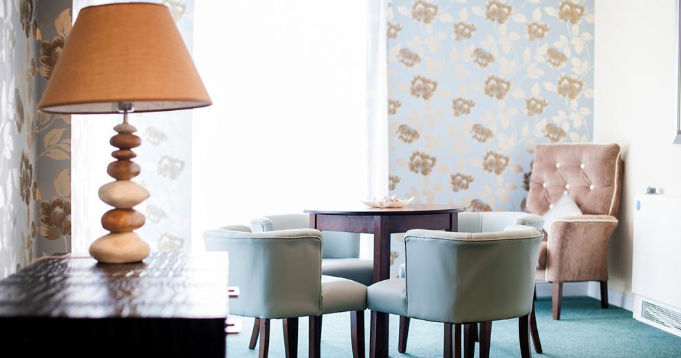 A wonderful airy seating area hosting an abundance of natural light from the large windows in the corners of the room. The pale blue wallpaper with earth toned floral patterns allows the light to reflect. The use of dark wooden furniture of chest of draws and circular tables break up the airy feel of the other lighter furniture such as four duck egg blue armchairs circling the table as well as a blushed pink crushed velvet chesterfield armchair facing towards the windows. The pop of orange in the lampshade but pairing it with the multi wood toned stoned design for the base.