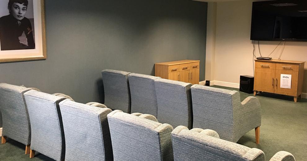 A theatre styled seating area, using greyish blue armchairs all facing towards a large wall mounted television, the dark green wall colour and dim lighting gives a atmospheric seating area.