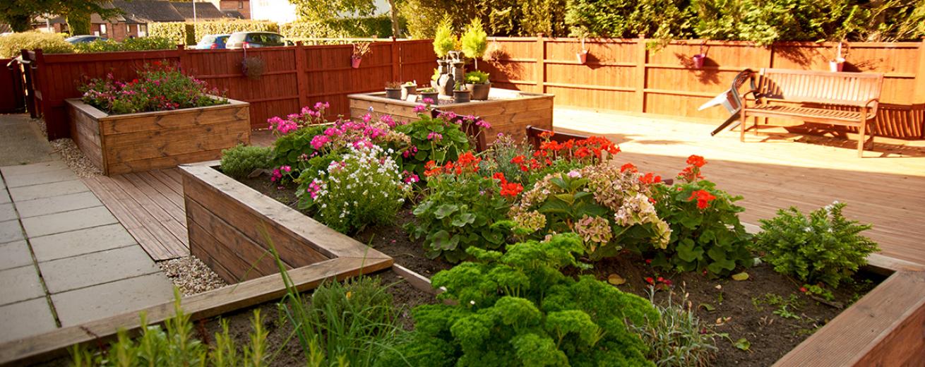 A low maintenance garden area, from a slab paved patio walkway onto the timber decking, which have been framed with a large wooden planter area and matching squared planters against the fencing, all featuring a beautiful array of vibrant flowers and unique pottery. There is also a large wooden bench to sit amongst the garden.