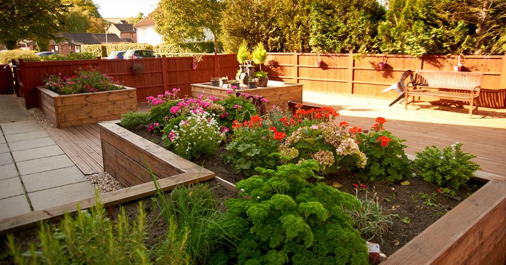 A low maintenance garden area, from a slab paved patio walkway onto the timber decking, which have been framed with a large wooden planter area and matching squared planters against the fencing, all featuring a beautiful array of vibrant flowers and unique pottery. There is also a large wooden bench to sit amongst the garden.