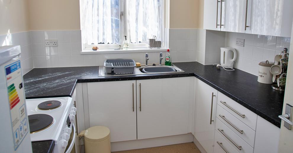 The brilliant white kitchen cupboards and stark contrast of the black mottled effect work top give the kitchen a clean and crisp feel. It hosts a white electric hob and oven, a fridge freezer and kettle.