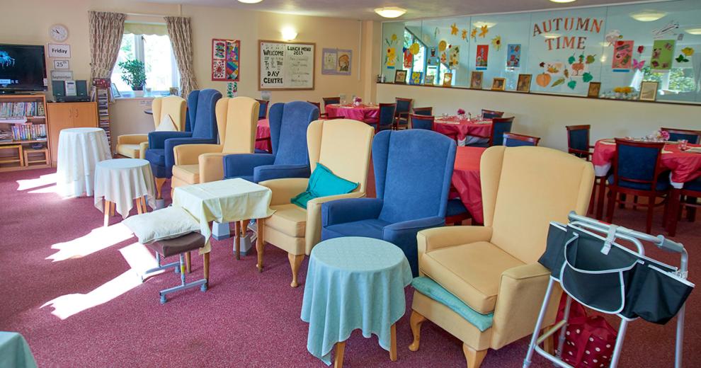 A large and brightly dressed communal seating area. The array of royal blue and yellow armchairs all face one way with a tall but petite table the sits just in front of the chairs one between two.