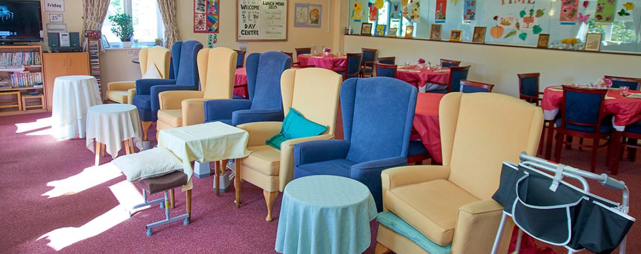 A large and brightly dressed communal seating area. The array of royal blue and yellow armchairs all face one way with a tall but petite table the sits just in front of the chairs one between two.