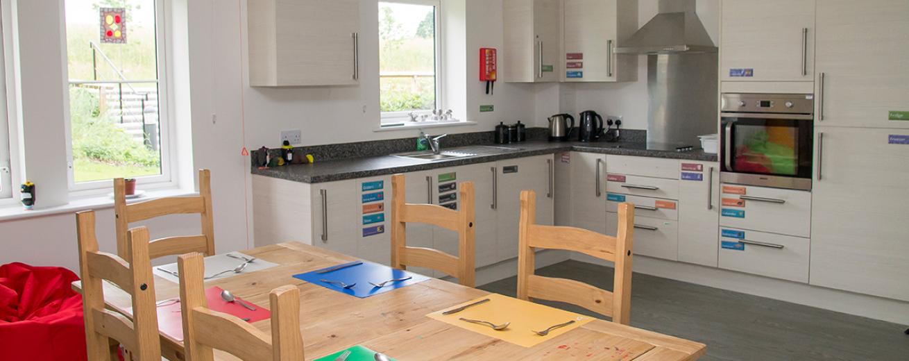 Bright corner unit kitchen with integrated appliances and colour coordinated stickers for accessible access to utensils. Standard pine wooden dining table with primary and secondary coloured place mats.