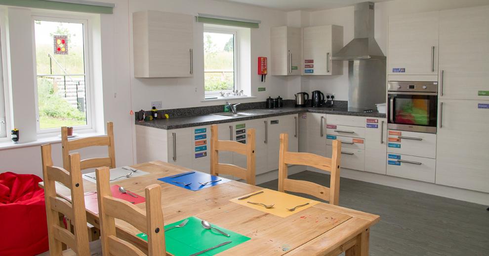 Bright corner unit kitchen with integrated appliances and colour coordinated stickers for accessible access to utensils. Standard pine wooden dining table with primary and secondary coloured place mats.