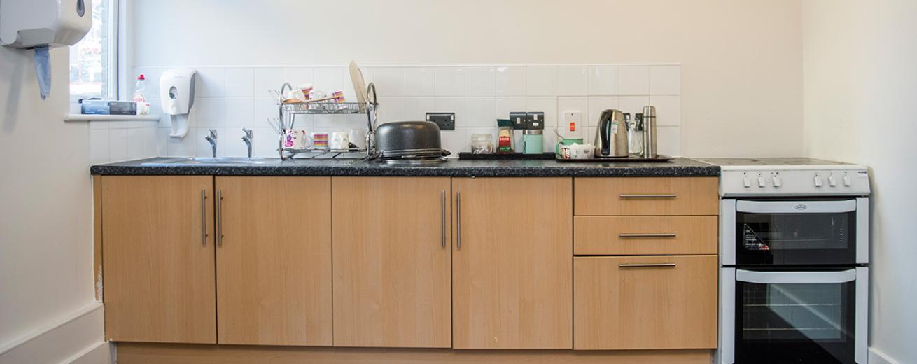 A communal kitchenette with two light wooden cupboards with three matching draws. A white electric oven and hob. The sink takes a large portion of the black mottled worktop, with a selection of tea making facilities to the right hand side of the sink.