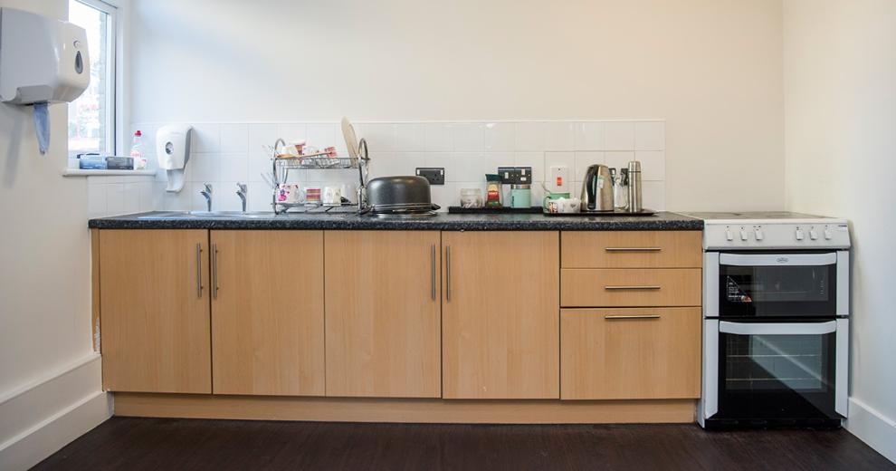 A communal kitchenette with two light wooden cupboards with three matching draws. A white electric oven and hob. The sink takes a large portion of the black mottled worktop, with a selection of tea making facilities to the right hand side of the sink.