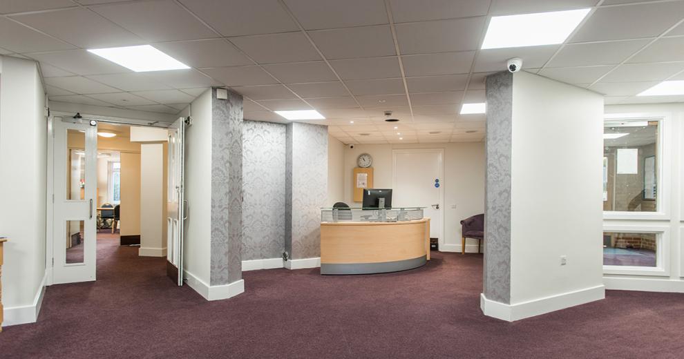 The reception area is tucked behind two partial walls which lead off into corridors. The soft silver fleur de lis wallpaper creates an inviting feel to the reception area which houses a curved corner desk with computer monitor.