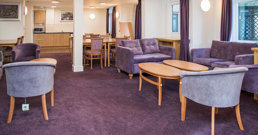 Multitoned purple seating area with plush lavender sofas, lilac bucket arm chairs surrounding soft wooden oval coffee tables and a kitchenette at the back of the room.