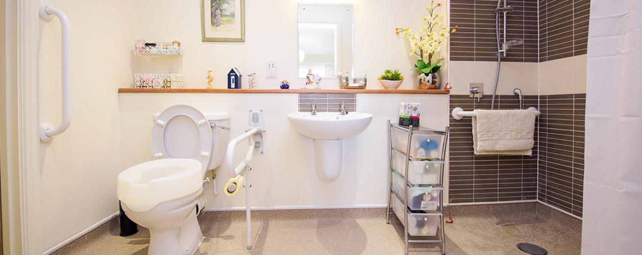 Well presented, sophisticated white painted and small grey rectangle tiled wet room with walk-in shower, white wall mounted sink and toilet. With a small shelf sitting in front of the wall mounted mirror hanging above the sink. The shelving is dressed in personal ornaments and potted plants to add a personal touch to the room.