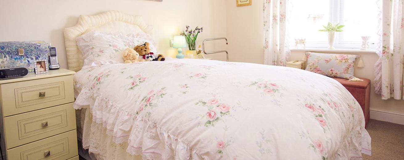 A serene single bedroom decorated in white tones. The frilly floral bedding adorned on a single bed. Adjacent are two bedside tables the left hand side features three draws. Under the window is a tan coloured ottoman providing seating and storage.