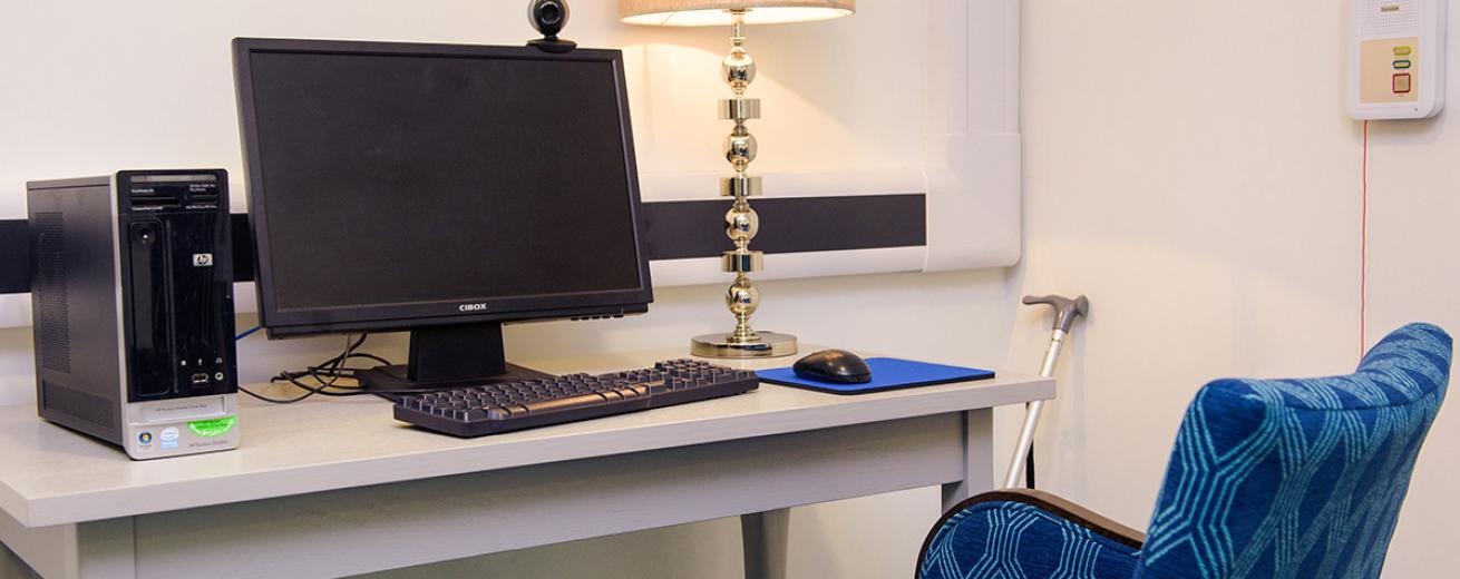 A modern computer area, the white wooden desk slots close to the corner of the room featuring an ornate metal desk lamp with pale pink lamp shade. The rest of the desk is filled with a modern computer monitor, computer tower, keyboard and mouse on a blue mouse mat. The blue links perfectly to the royal blue arm chair with a light blue geometric print.