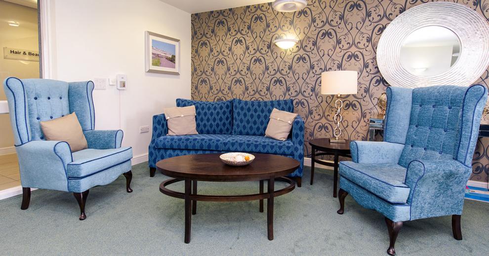 A sophisticated communal seating area. Displaying a bronzed base colour with a navy swirl repeat patterned design feature wall, against the white painted walls. The dark wooden oval coffee table and circular side table complement the navy two seater sofa and two comfy light blue chesterfield armchairs.