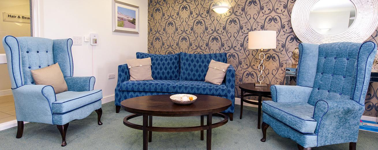 A sophisticated communal seating area. Displaying a bronzed base colour with a navy swirl repeat patterned design feature wall, against the white painted walls. The dark wooden oval coffee table and circular side table complement the navy two seater sofa and two comfy light blue chesterfield armchairs.