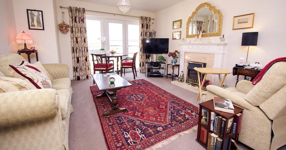 A fair sized comfortable lounge, a large two seater cream sofa faces the electric fire, within a white wooden surround. Above the fire a scalloped effect wooden mirror fills the space with framed photographs hung on the wall. A modern television on top of black and silver television unit tuck into the corner, next to a small dark wooden table with a floral display. In front of the patio doors the small two person dining room table over looks the views. An oversized red patterned rug breaks the room up and adds a vibrant pop of colour.