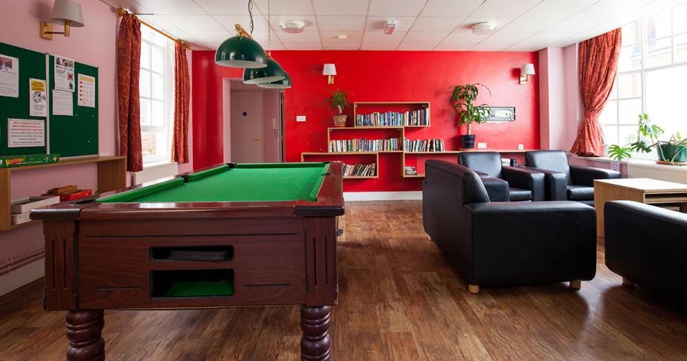 Bold red communal area, wall shelving holding numerous books and potted plants. Numerous leather seating options next to a grand pool table and notice boards.