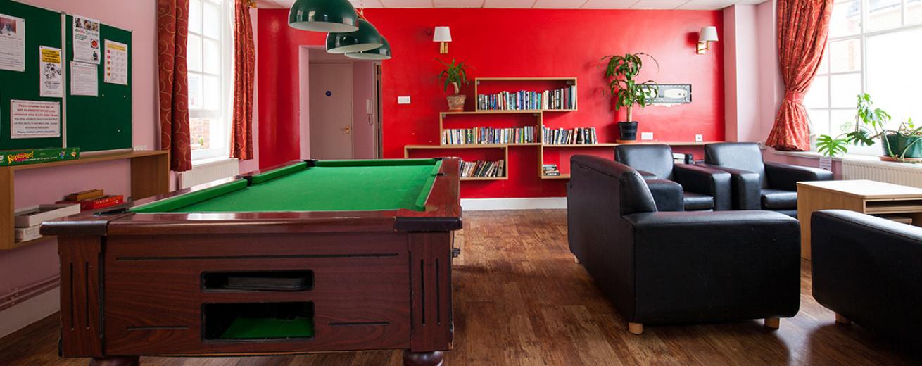 Bold red communal area, wall shelving holding numerous books and potted plants. Numerous leather seating options next to a grand pool table and notice boards.