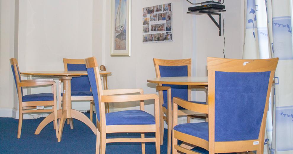 Blue upholstered wooden dining room chairs placed around two small circular tables with sea view and resident photographs hung on the walls.