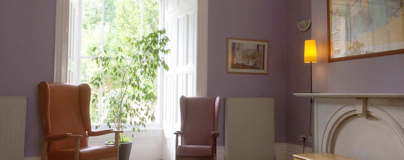 Serene purple space with a central alcove leading to a sizeable window, decorated with panelling and home to an indoor tree plant. Also featuring two grand armchairs either side.